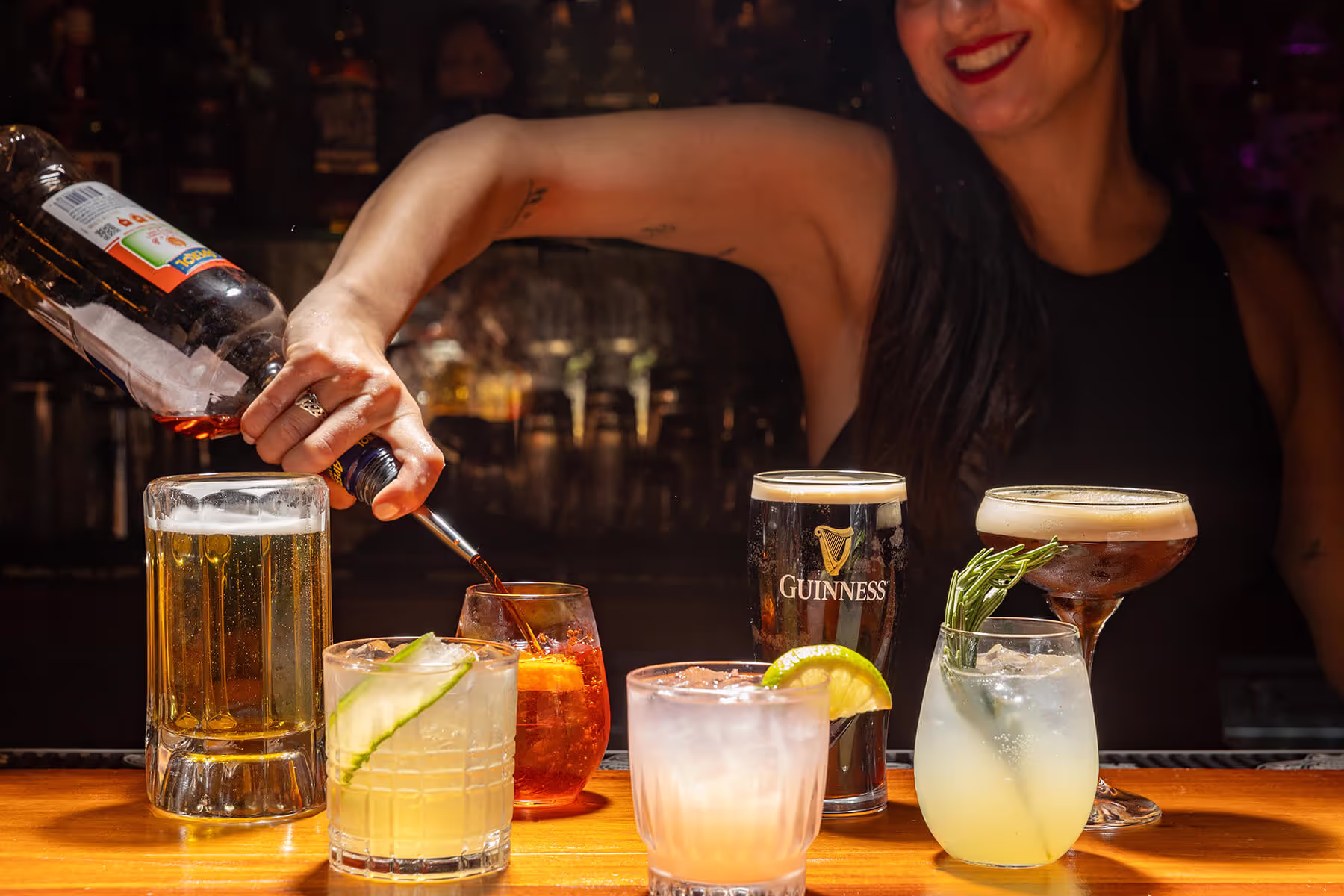 Bartender pouring a drink behind a wooden bar with an assortment of cocktails and a pint of Guinness in front.