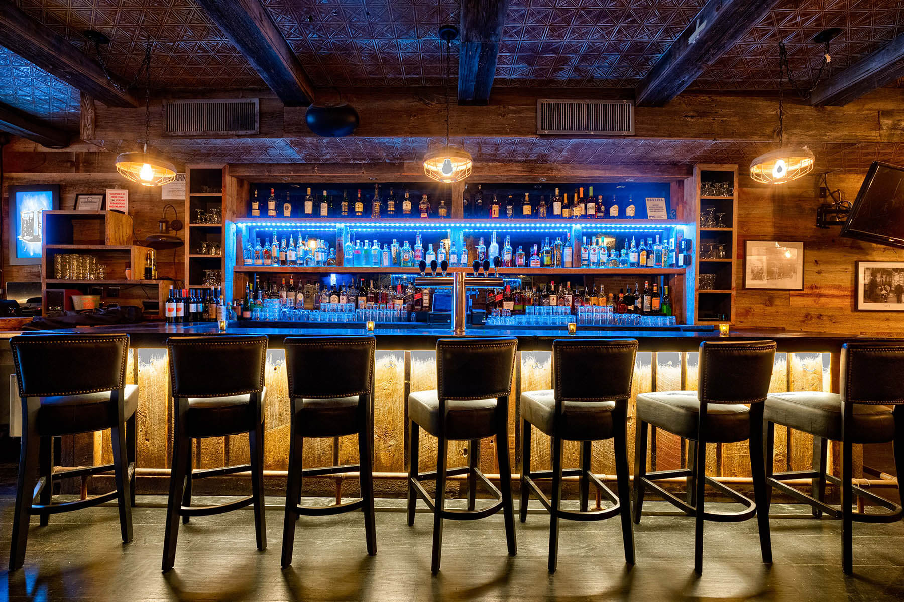 Wood-themed bar interior with high chairs lined up under a counter illuminated by warm lighting and blue backlit shelves stocked with bottles.